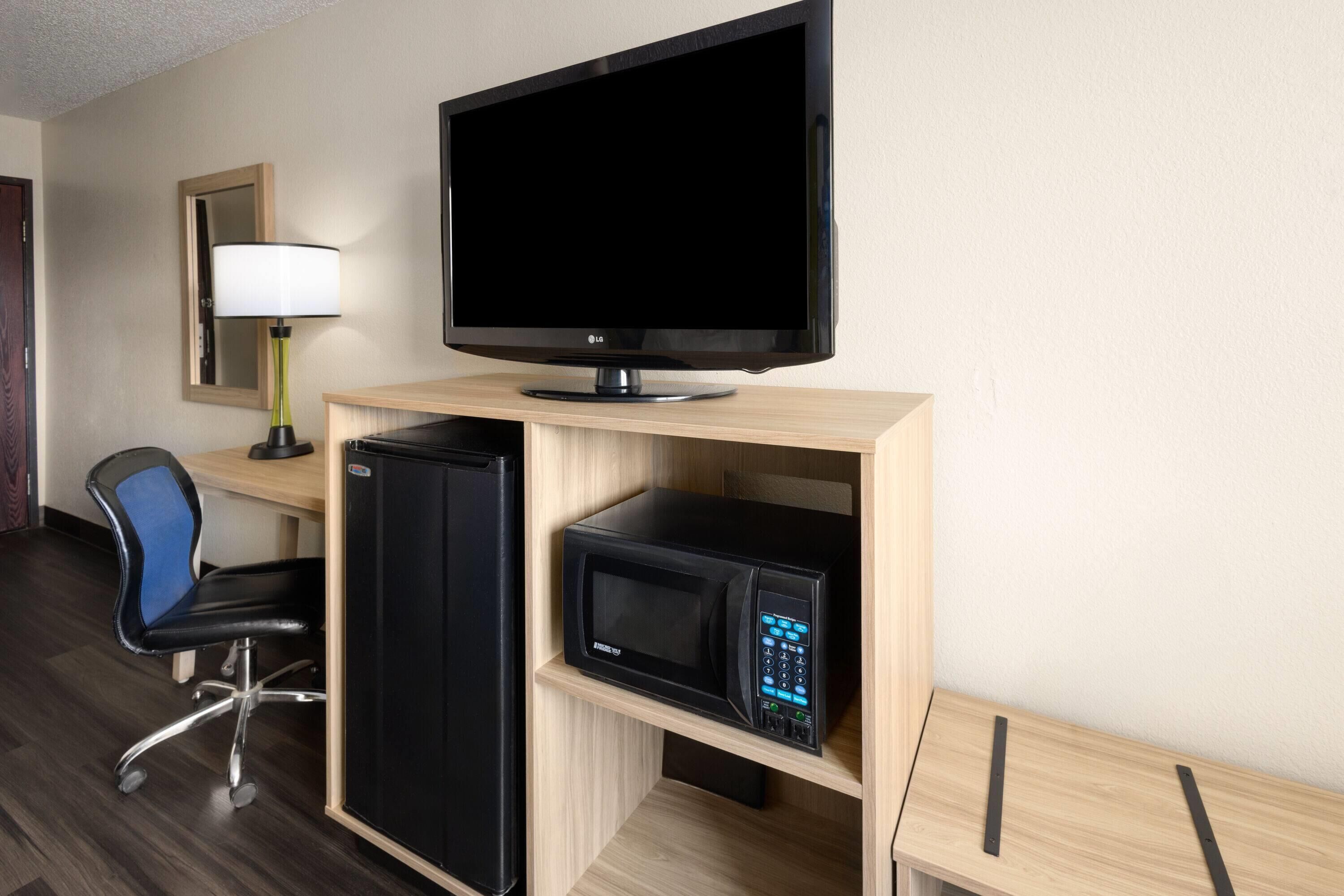 A hotel room with a flat-screen TV, a black refrigerator, a microwave, a desk with a chair, and a lamp.