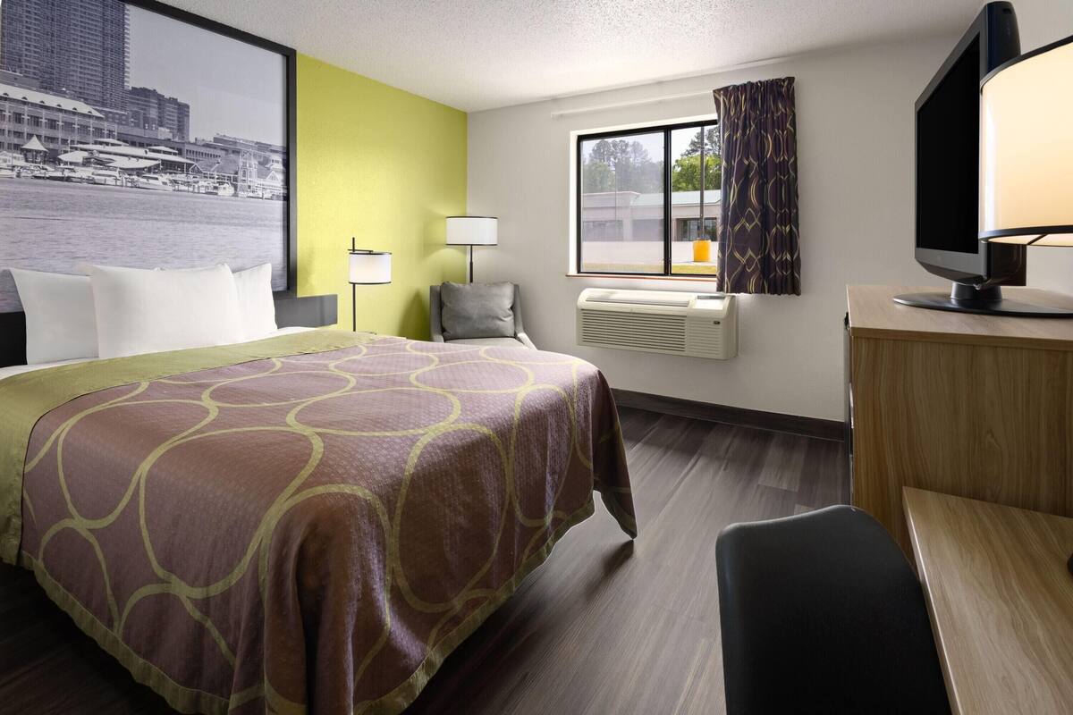 Habitación de hotel con cama, escritorio, televisión, ventana con cortinas y aire acondicionado.