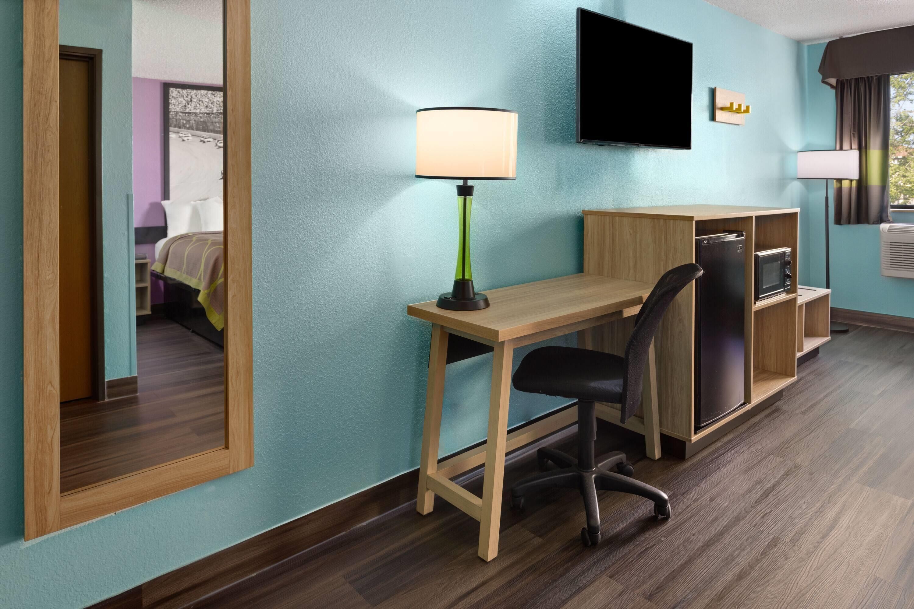 A hotel room with a wooden desk, a black office chair, a television mounted on the wall, and a window with curtains.
