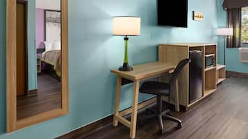 A hotel room with a wooden desk, a black office chair, a television mounted on the wall, and a window with curtains.