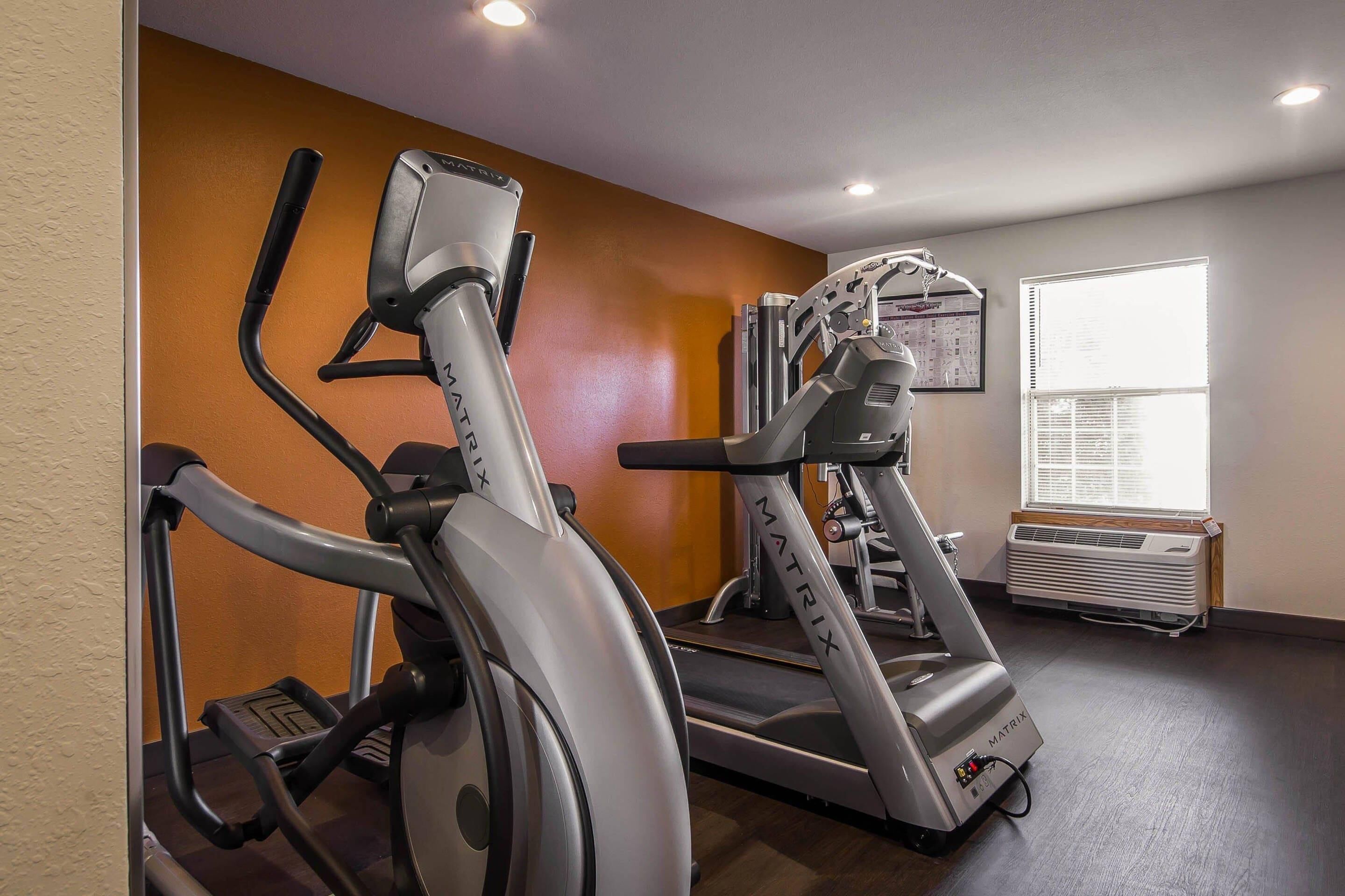 Un gimnasio con bicicletas estáticas y cintas de correr, una ventana con persianas y un equipo de aire acondicionado instalado en la pared.
