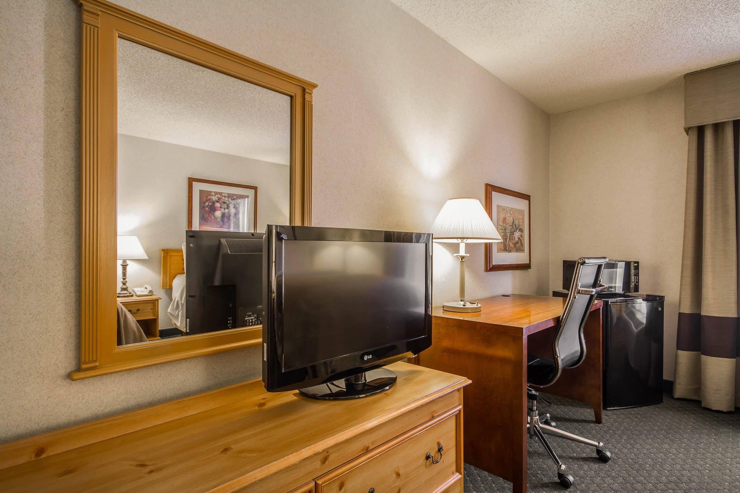 A hotel room with a wooden desk, a television, a lamp, a chair, a mirror, and a microwave.