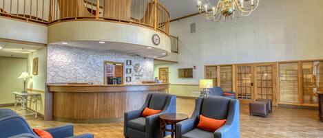 A hotel lobby with a reception desk, two armchairs with orange cushions, a chandelier, and a wooden staircase.