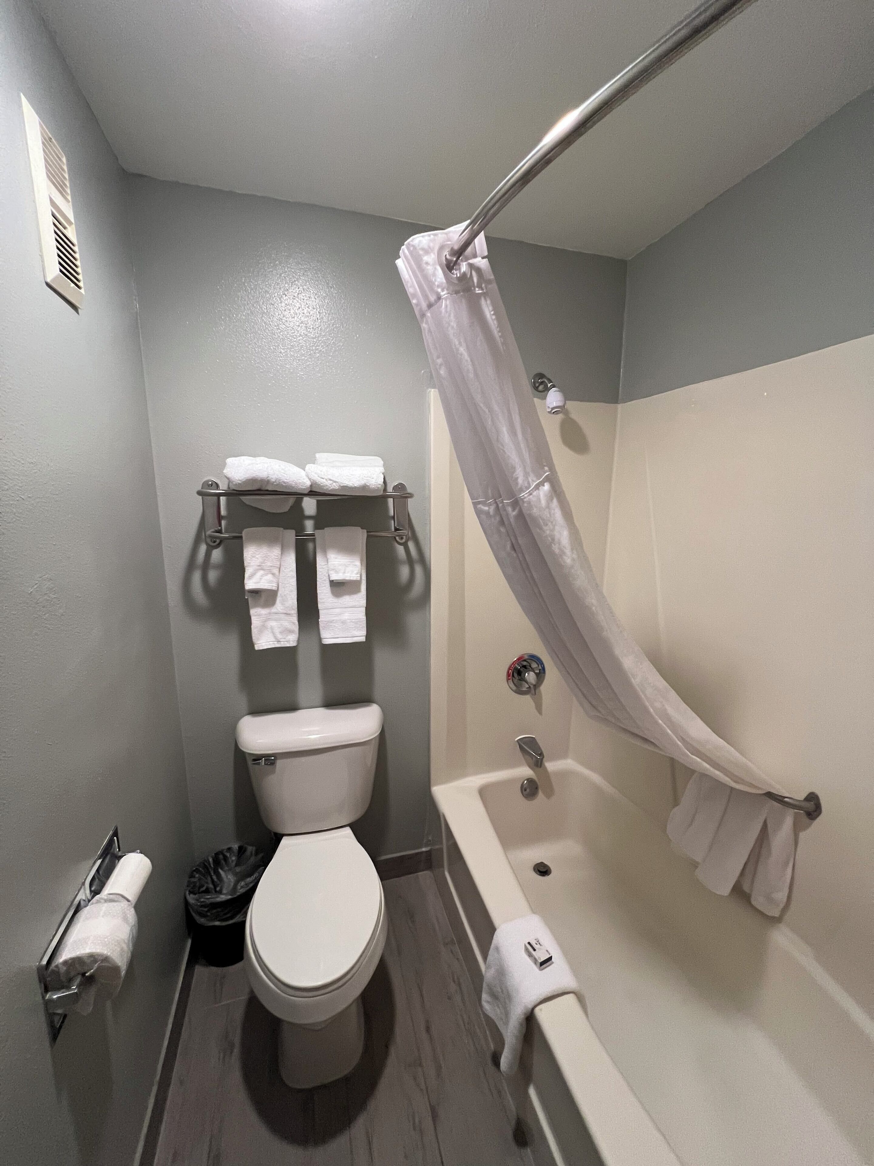 A small bathroom with a toilet, a shower with a curtain, and a towel rack with multiple towels.