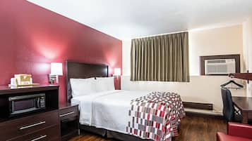 Hotelkamer met een bed, een bureau met een computer, een magnetron, een rode stoel en een raam met gordijnen.