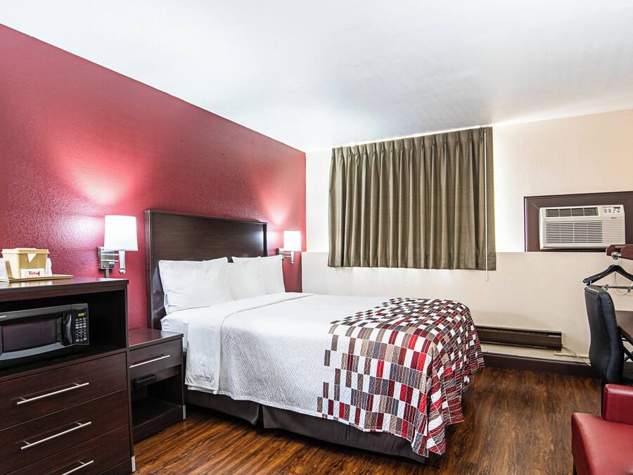 A hotel room with a bed, a desk with a computer, a microwave, a red chair, and a window with curtains.