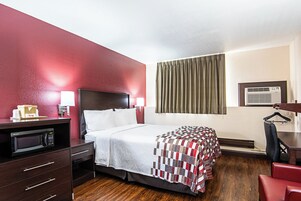 Habitación de hotel con cama, escritorio con computadora, microondas, una silla roja y ventana con cortinas.
