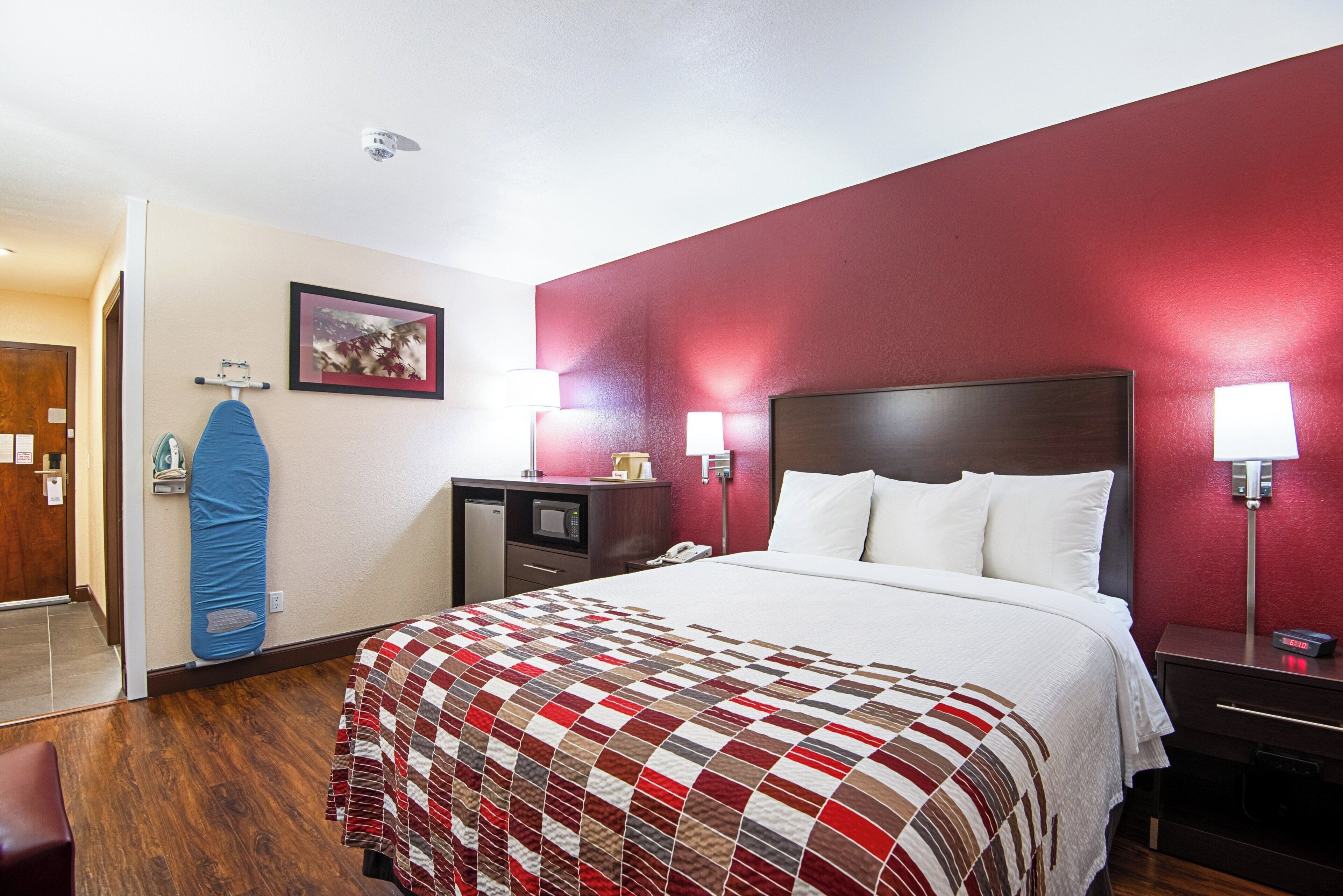 A hotel room with a bed, a desk, a microwave, a telephone, and a wall-mounted lamp.