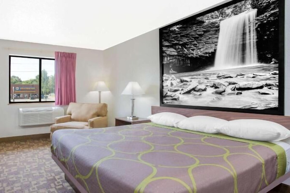 Habitación de hotel con cama, escritorio, silla, ventana con vistas y un cuadro de una cascada colgado en la pared.