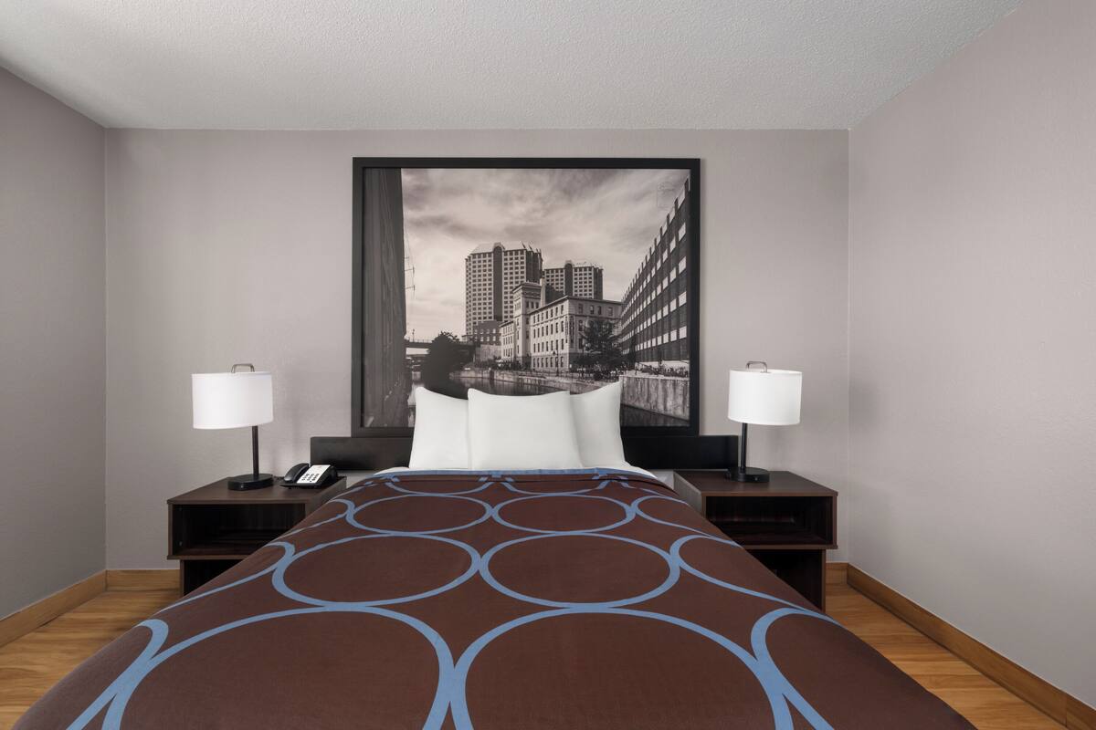 A hotel room with a bed, two bedside tables, a nightstand, a telephone, and a large framed cityscape picture on the wall.