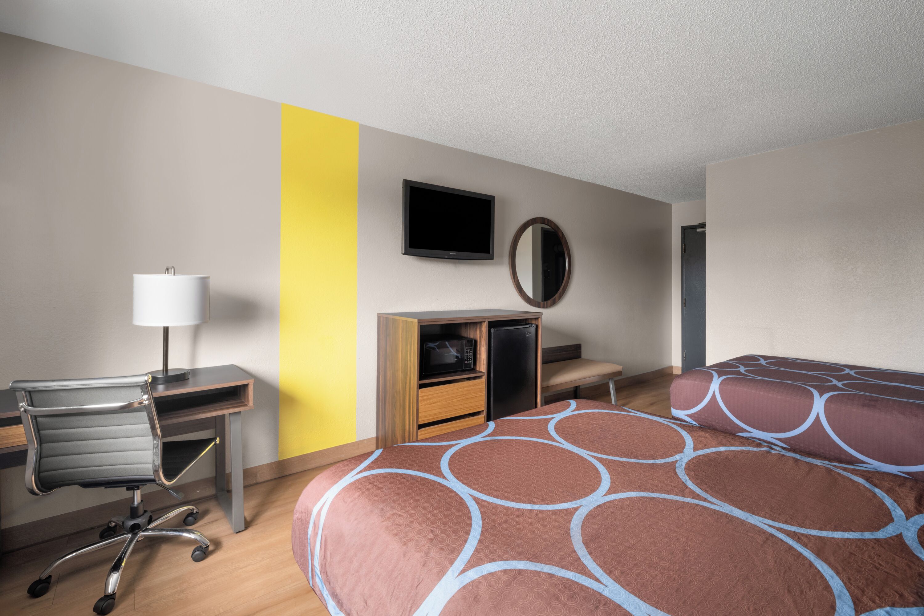 A hotel room with a desk, chair, bed, TV, and a mirror.