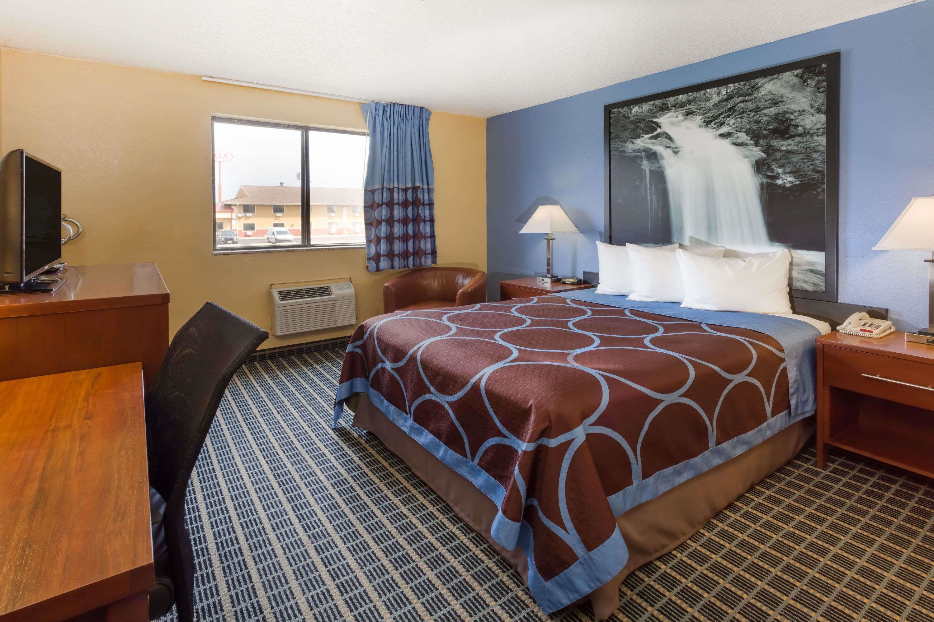 Una habitación de hotel con cama, escritorio, silla, televisor, ventana con vistas a edificios y un cuadro grande con una cascada.