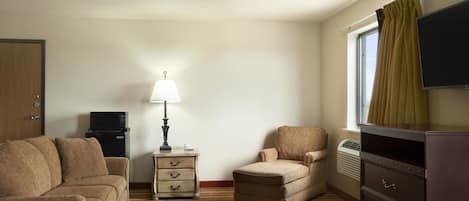 A hotel room with a sofa, armchair, dresser, lamp, TV, and a window with curtains.
