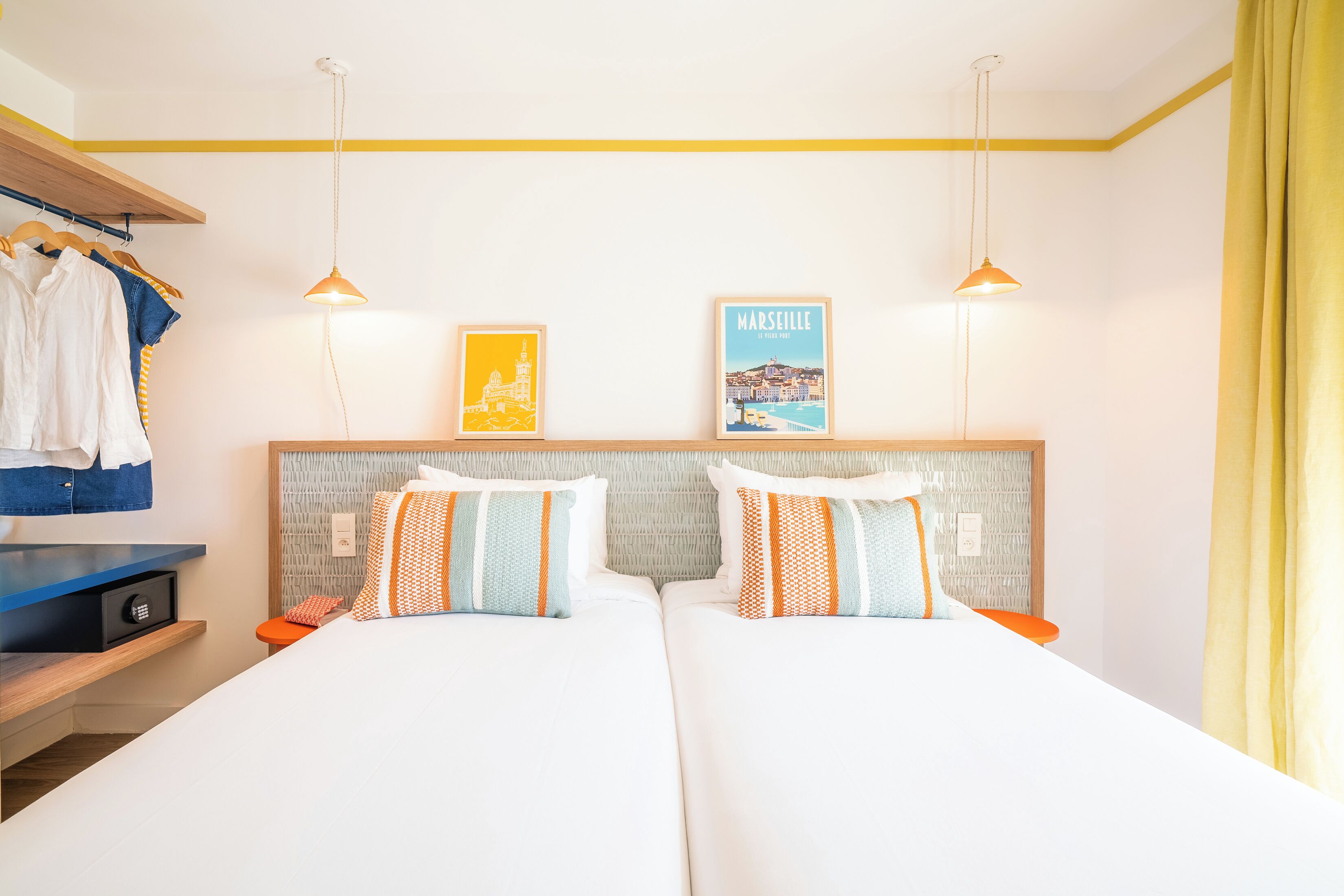 A hotel room with two beds, a wardrobe, a desk, and a wall with two framed pictures.