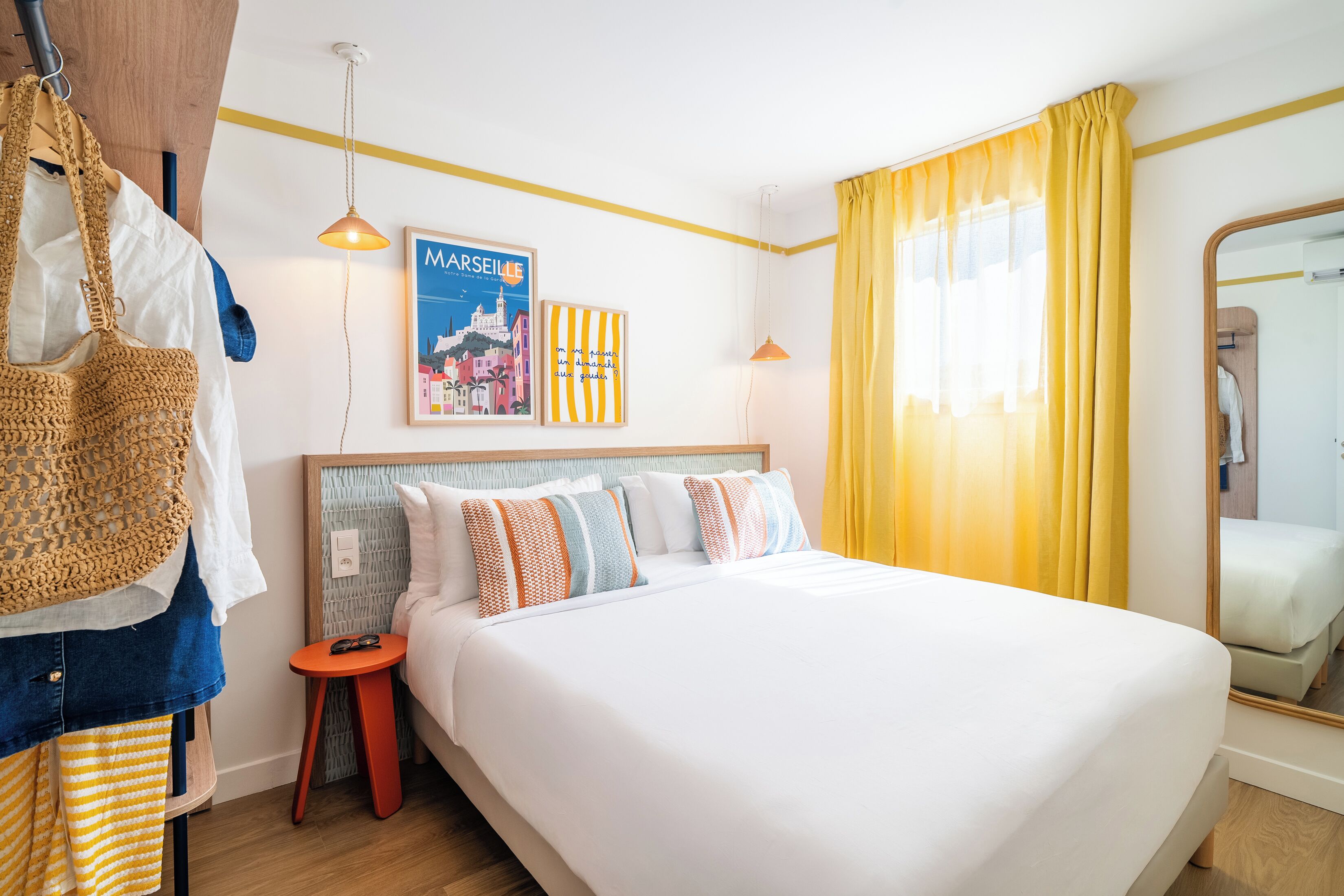 A hotel room with a bed, a headboard, a nightstand, a hanging bag, a framed picture, and a mirror.