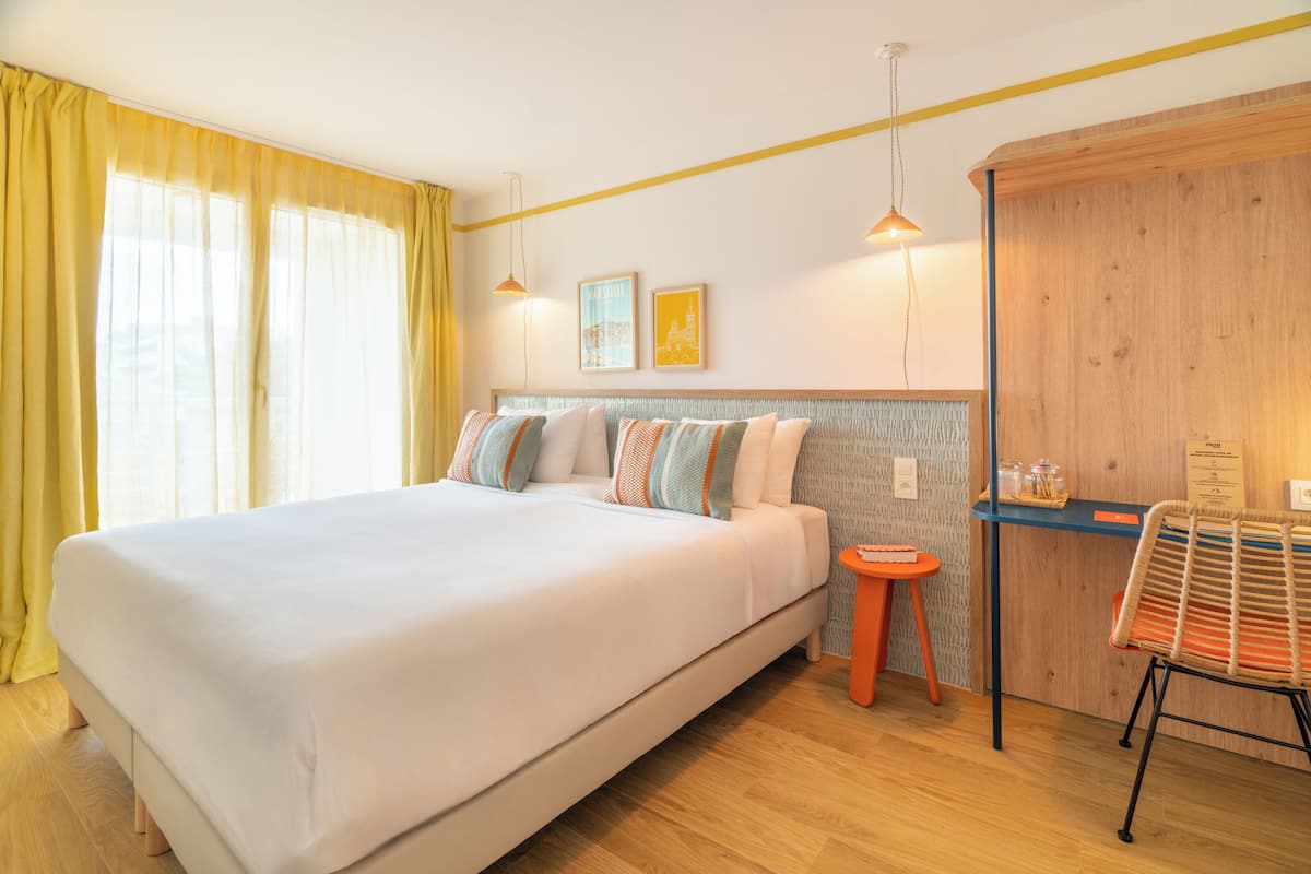A hotel room with a bed, a small table, a chair, and a wooden wardrobe.