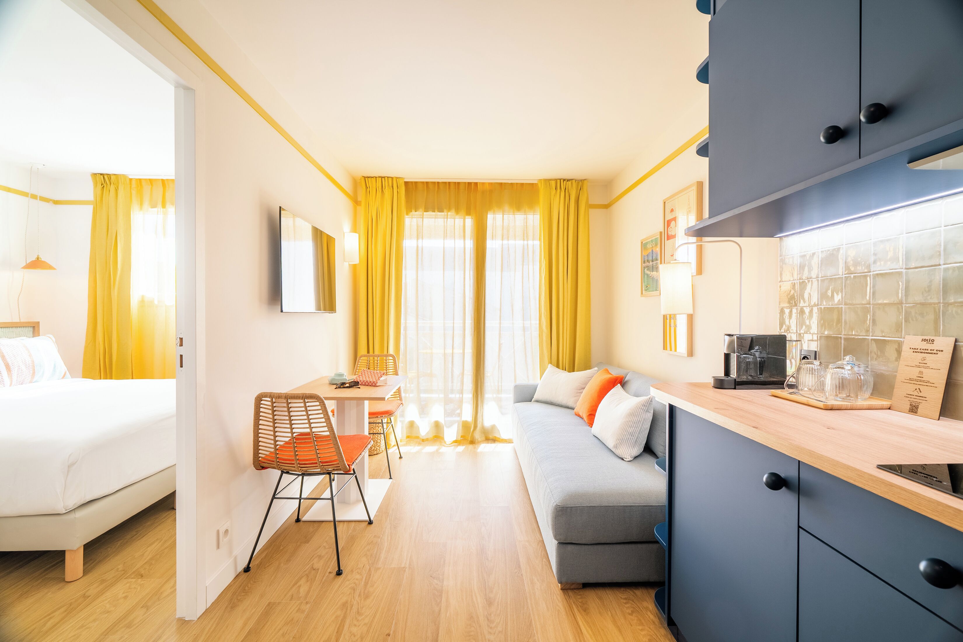 A compact hotel room with a bed, a sofa, a dining area, and a kitchenette.