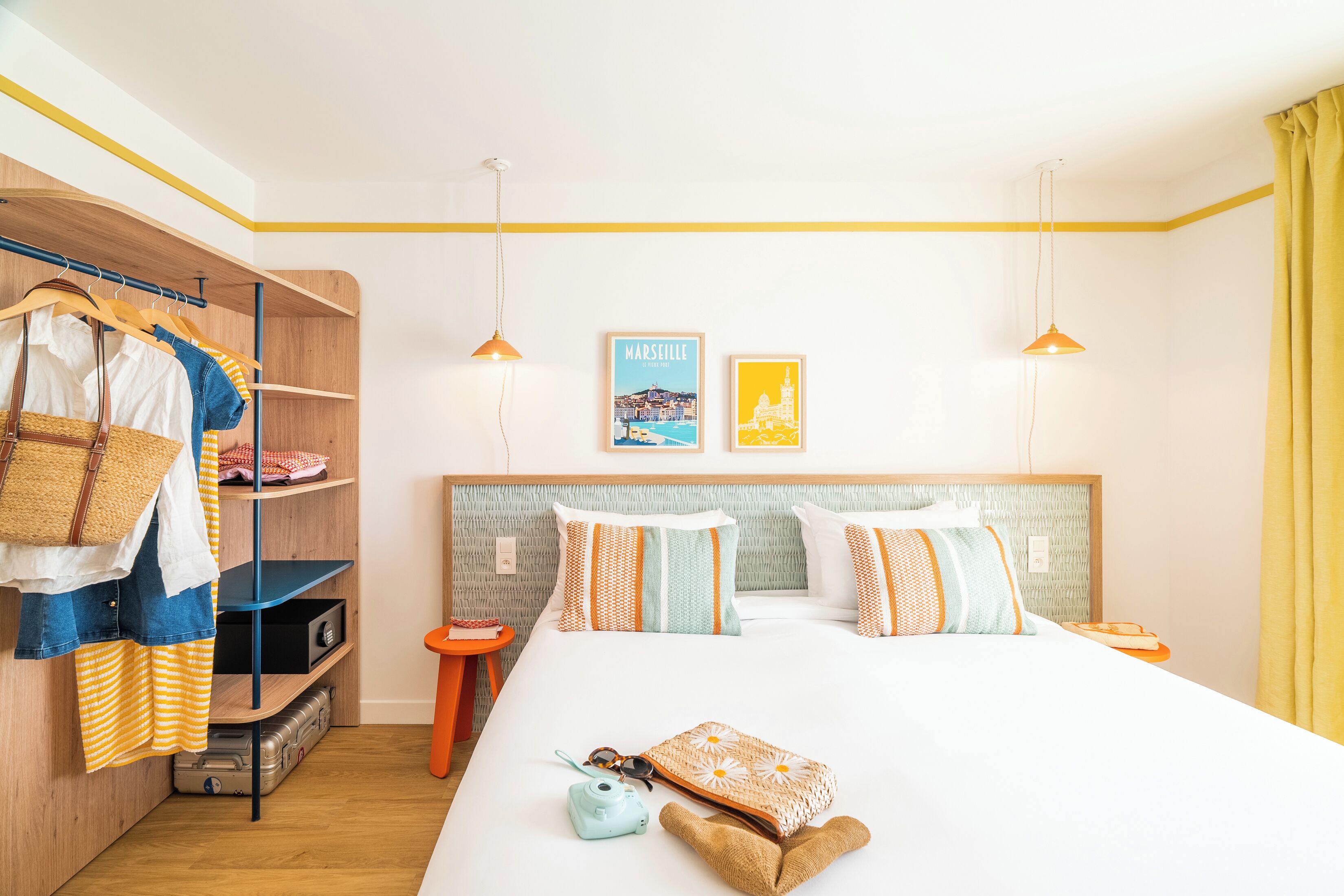 Hotelkamer met een bed, een kledingkast, een plank met kleding, een klein tafeltje en twee aan de muur hangende foto's.