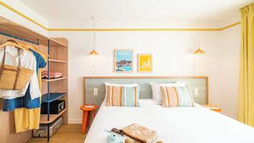 Une chambre d’hôtel comprenant un lit, une armoire, une étagère avec des vêtements, une petite table et deux tableaux accrochés au mur.