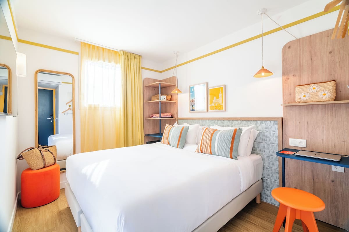 A neatly arranged hotel room with a large bed, a wooden headboard, a small desk, and a stool.