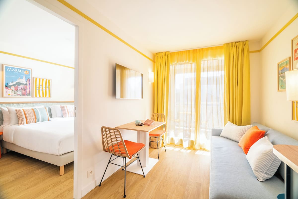 A hotel room with a bed, a sofa, a dining table with chairs, a TV, and a window with curtains.