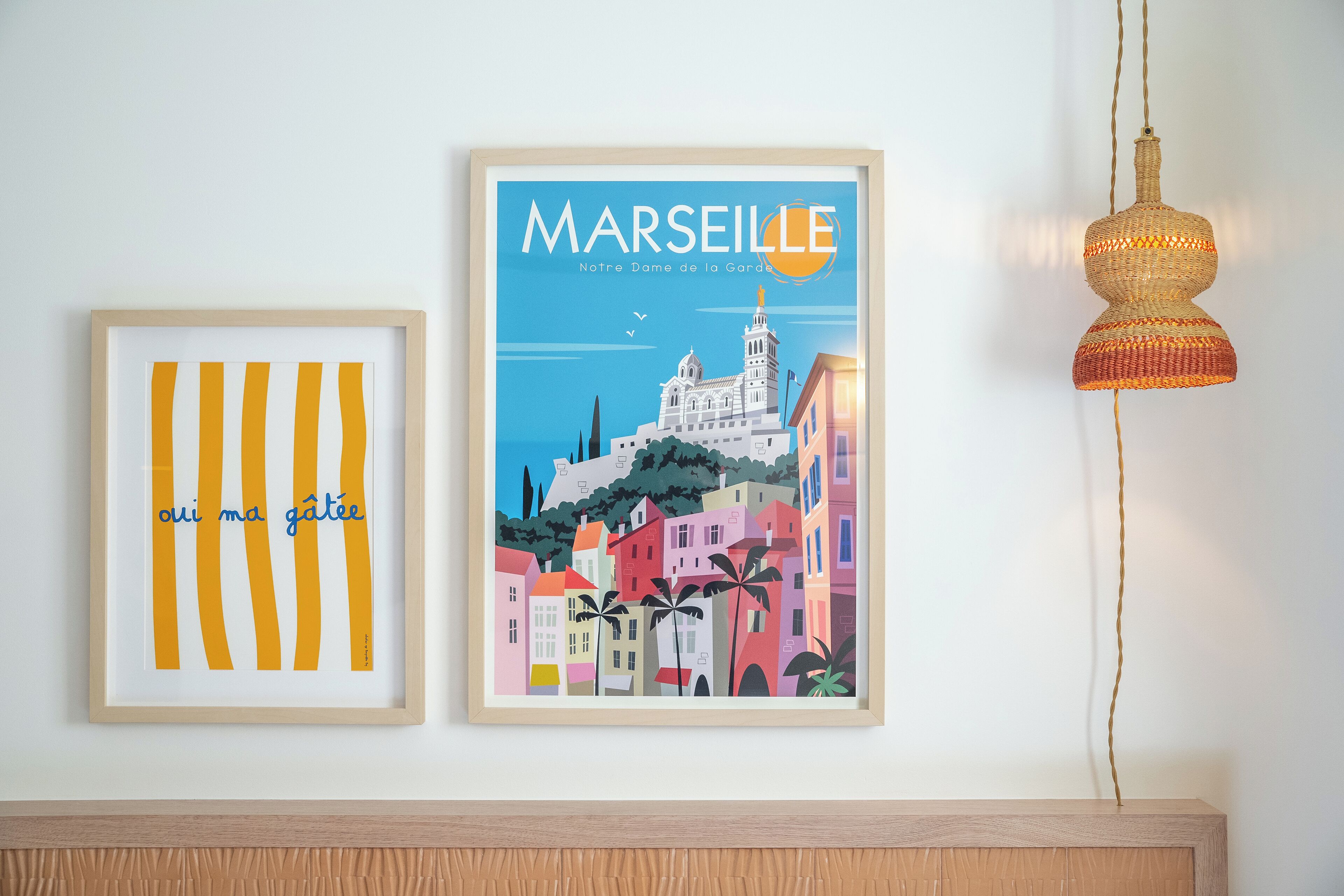 Two framed artworks on a wall, one depicting a colorful cityscape with the text "MARSEILLE Notre Dame de la Garde" and the other with vertical yellow stripes and the text "oui ma gatée".
