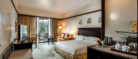 Habitación de hotel con cama, escritorio con televisor, silla, mesa con lámpara y vistas al exterior.