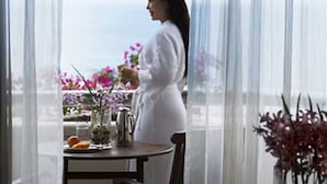 Eine Person in einem weißen Bademantel steht an einem Tisch mit einer Kanne und Blumen, mit Blick auf eine schöne Aussicht.