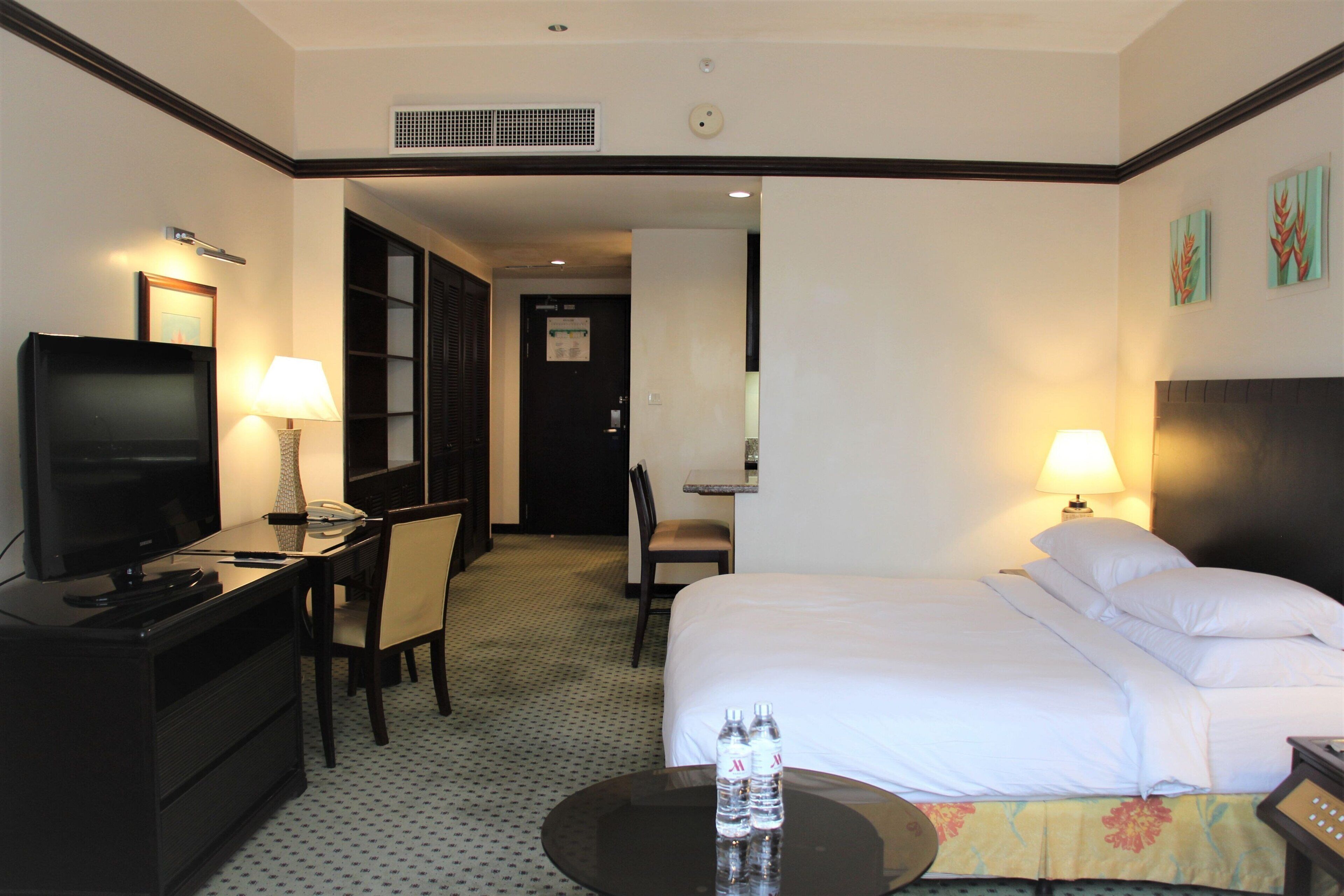 A hotel room with a bed, a desk with a television, a chair, a table with two water bottles, and a lamp.