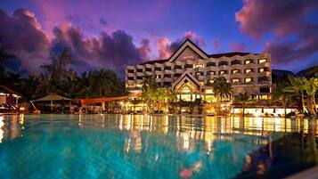 A hotel with a swimming pool, palm trees, and a sunset sky.