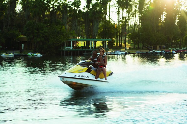 A watercraft with two people on it, moving on a body of water with trees and buildings in the background.