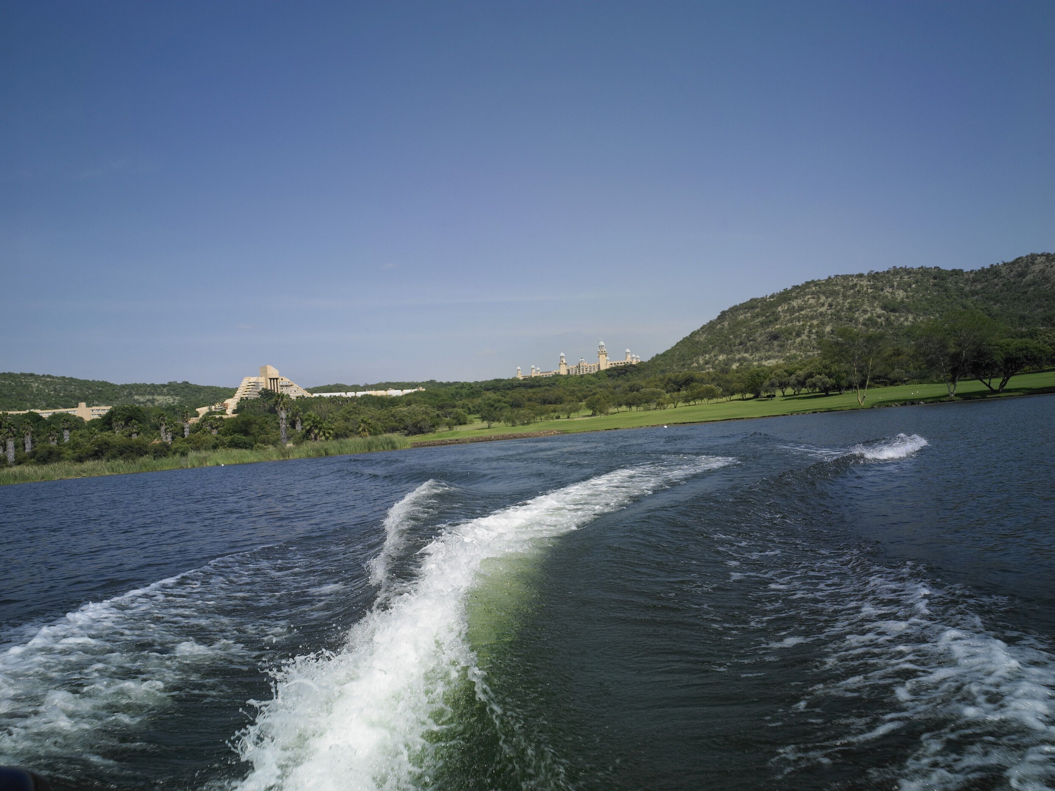 A body of water with a wake behind a moving boat, a green landscape with trees, and a distant building with multiple towers.