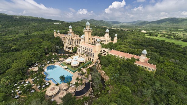A large, historic building with multiple towers and domes, surrounded by lush greenery and a swimming pool. - The Palace of the Lost City at Sun City Resort (Moses Kotane)