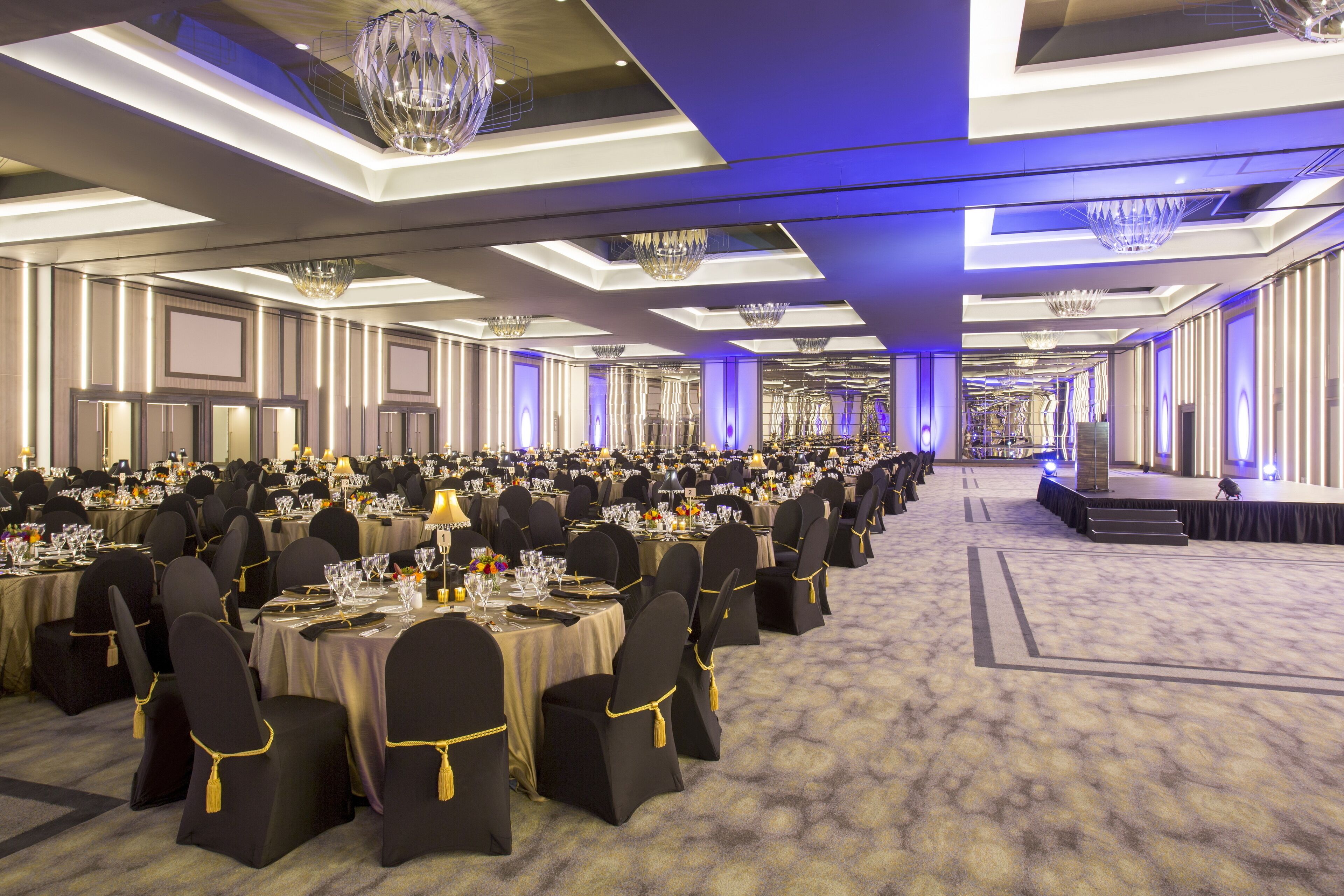 A banquet hall with rows of round tables, black chairs with gold accents, and a stage at the front.