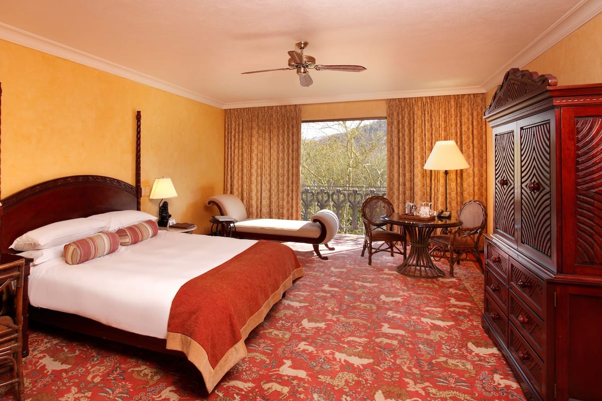 A hotel room with a large bed, two chairs, a table, a TV, and a balcony with curtains.