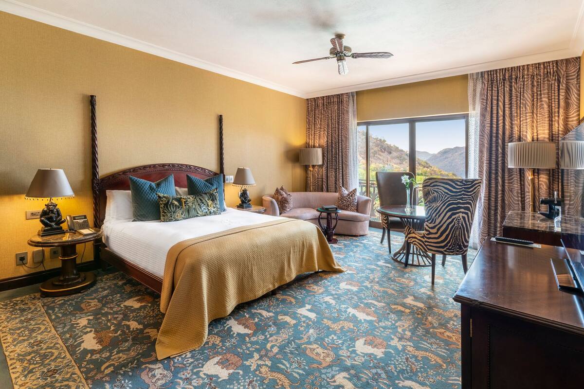 A hotel room with a bed, a desk, a chair, and a sofa. There is a view of the outdoors through the window.