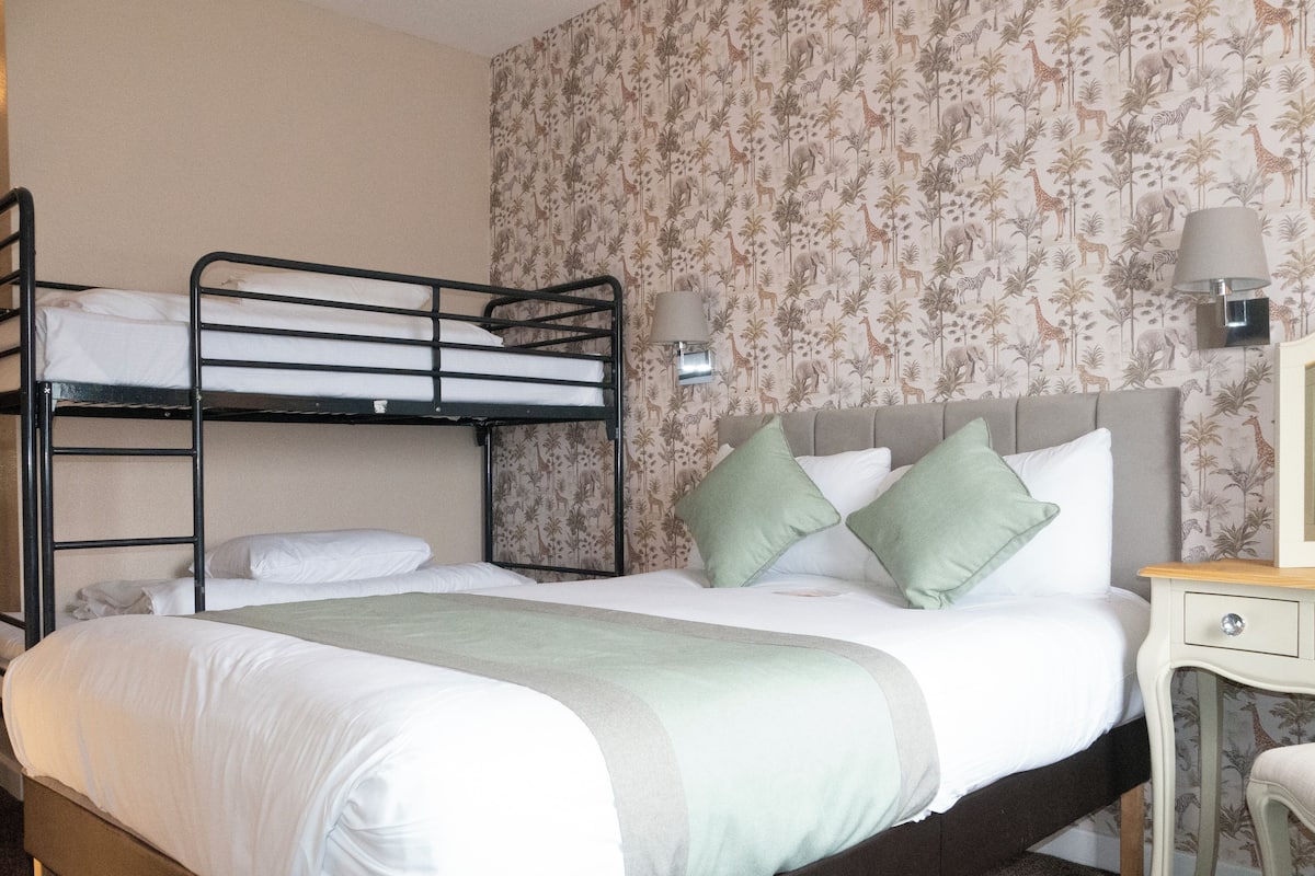 A bunk bed room with a single bed, green pillows, a bedside table with a lamp, and a wall with animal wallpaper.