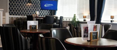 A hotel lobby with a television displaying the Capital logo, multiple tables and chairs, and a patterned wall.