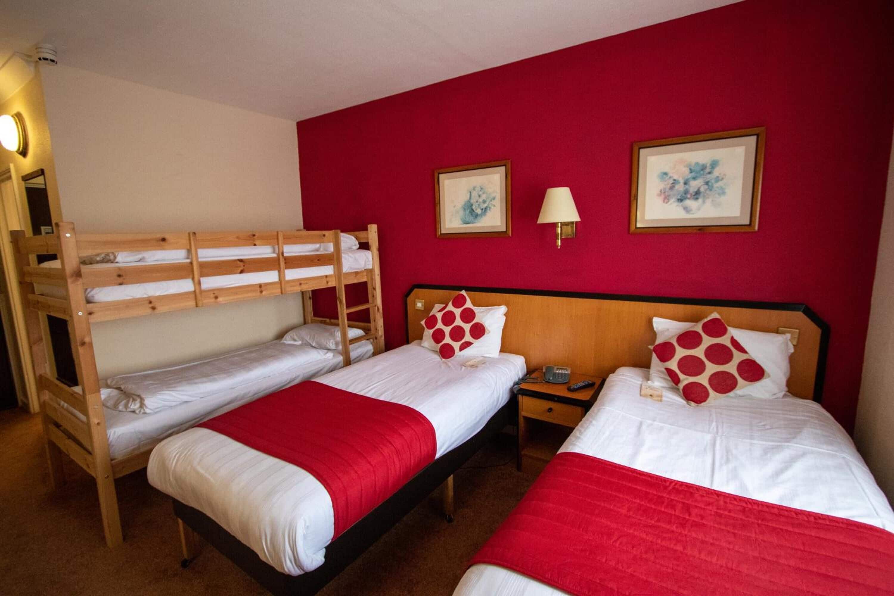 A room with two single beds and a bunk bed, all with white bedding and red accents. There are two framed artworks on the red wall, a bedside lamp, and a small table with a lamp.