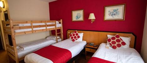A room with two single beds and a bunk bed, all with white bedding and red accents. There are two framed artworks on the red wall, a bedside lamp, and a small table with a lamp.