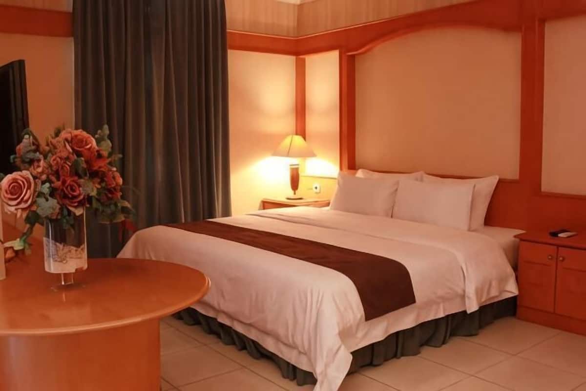 A hotel room with a bed, a bedside table with a lamp, a vase with flowers, and a round table.