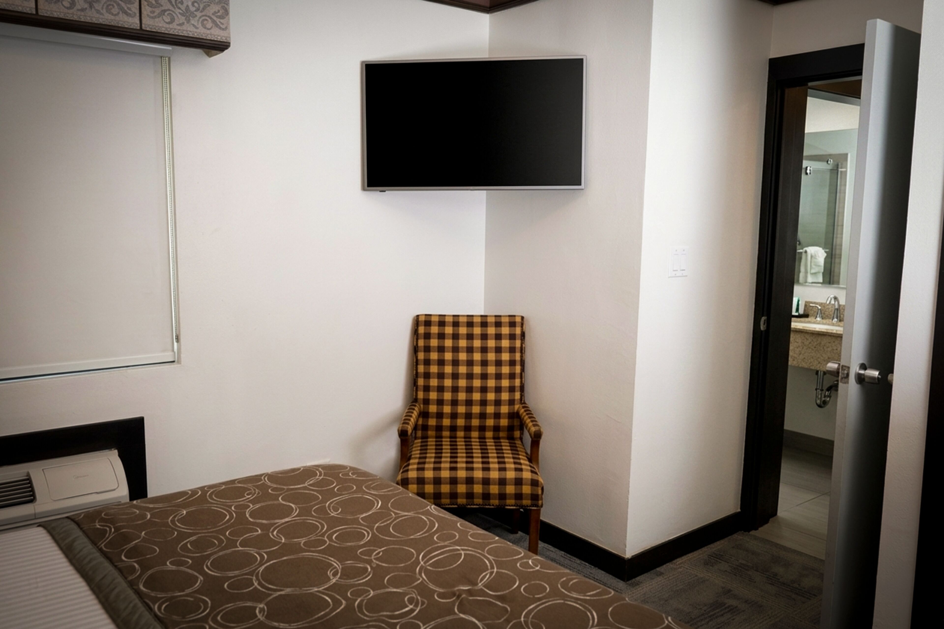 Habitación de hotel con cama, un sillón a cuadros, un televisor de pantalla plana en la pared y una puerta que da a un baño.