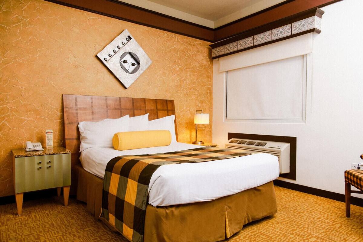 A hotel room with a bed, a nightstand, a lamp, a window with blinds, and a wall-mounted phone.