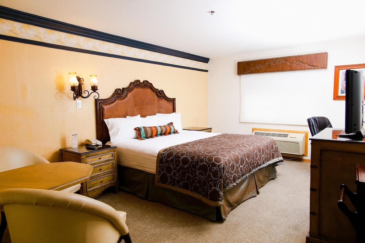 A hotel room with a wooden headboard bed, a desk with a chair, a bedside table, a lamp, and a television.