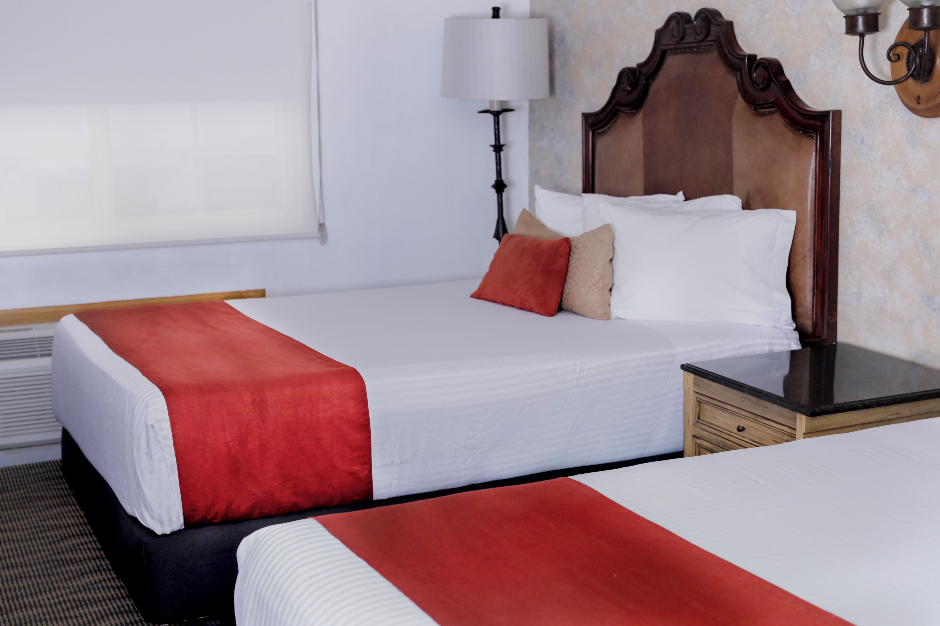 A hotel room with two beds, a nightstand, a lamp, and a wall-mounted light fixture.