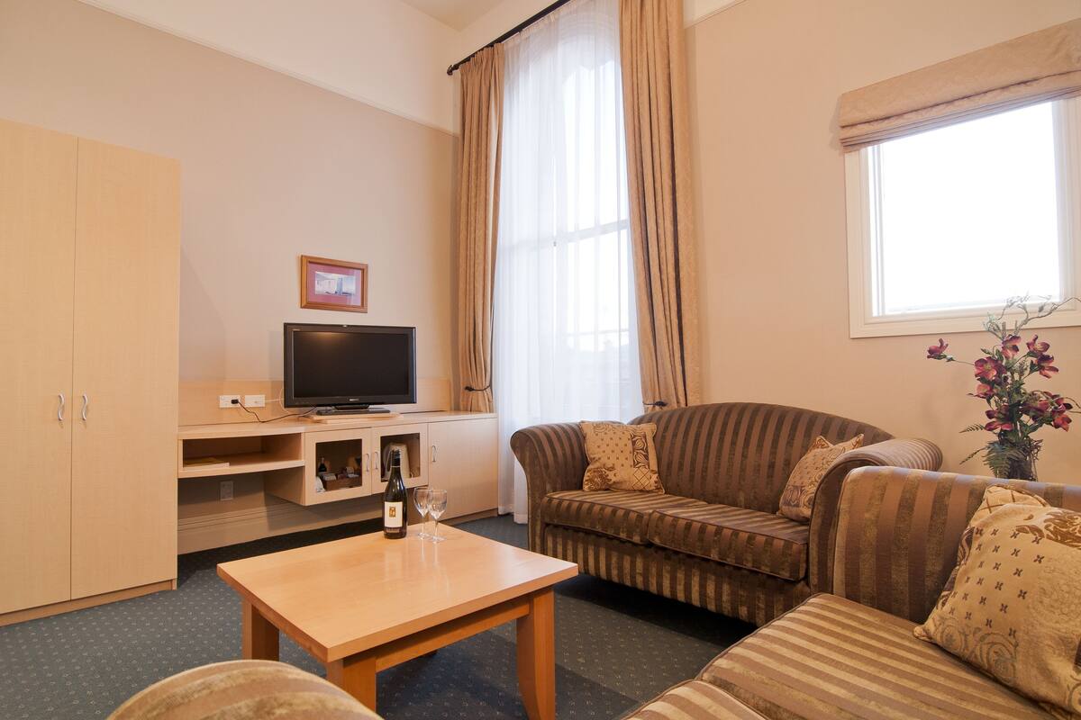Uma sala de estar com sofá, televisão, mesa de madeira e janela com cortinas.