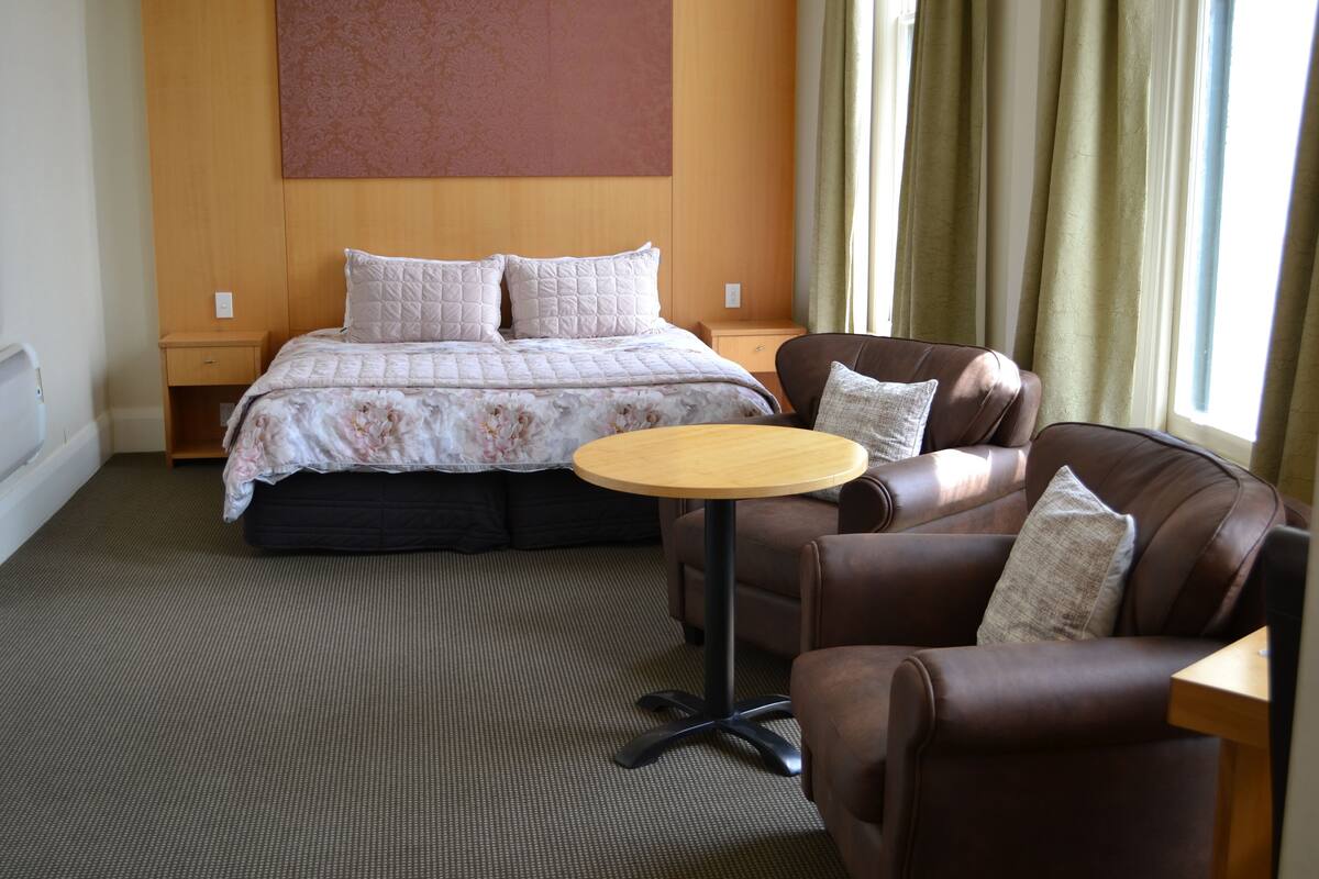 Habitación de hotel con cama, sofá de cuero marrón, una mesita redonda y una ventana con cortinas.