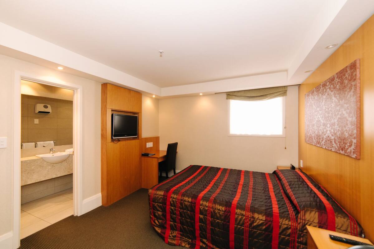 Habitación de hotel con cama, escritorio, televisión, baño con lavamanos y espejo, y ventana.