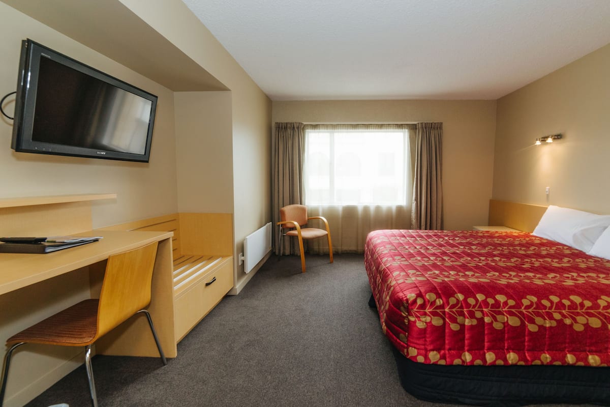 Habitación de hotel con cama, escritorio, silla, televisor y ventana con cortinas.