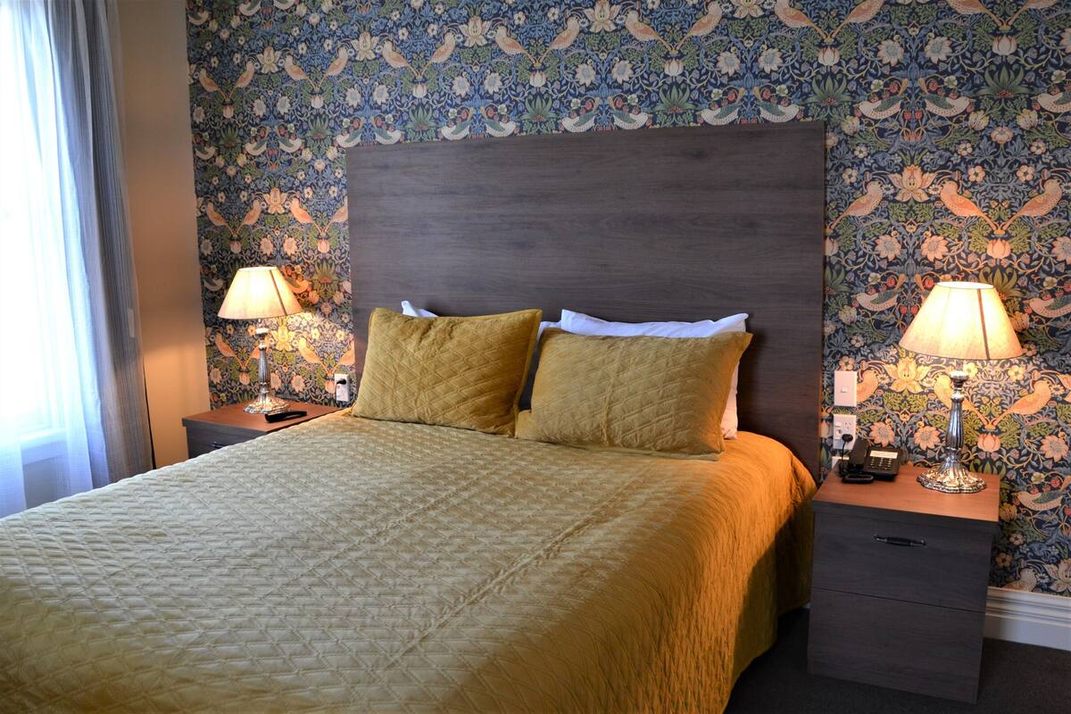 Cama bem arrumada com cobertura acolchoada, duas luminárias de cabeceira e mesa de cabeceira com telefone e abajur.